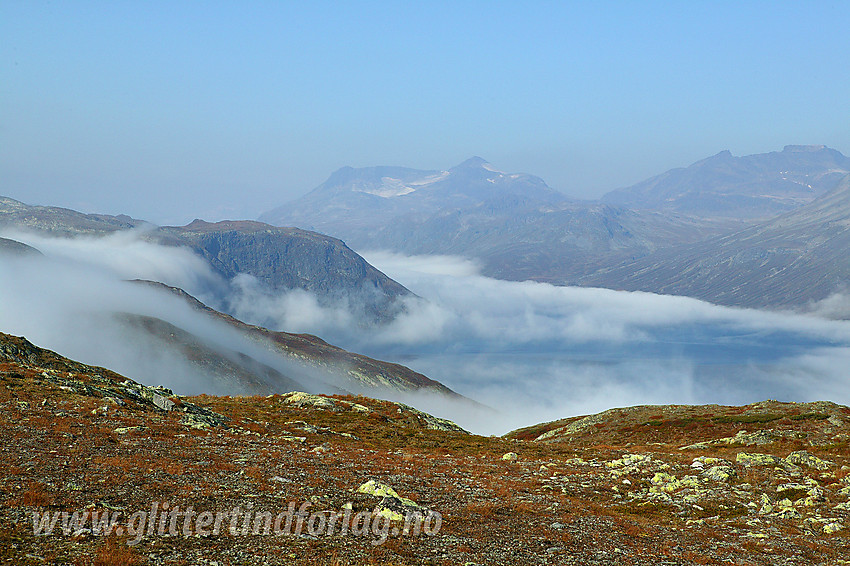 Tåkeskyer, nesten som scenerøyk, kommer veltende over fjellryggen fra sør, samtidig som den siver bortetter Bygdins vannflate. I den disige bakgrunnen ses bl.a. Galdebergtinden (2075 moh), Slettmarkpiggen (2164 moh) og Slettmarkhøe (2190 moh).