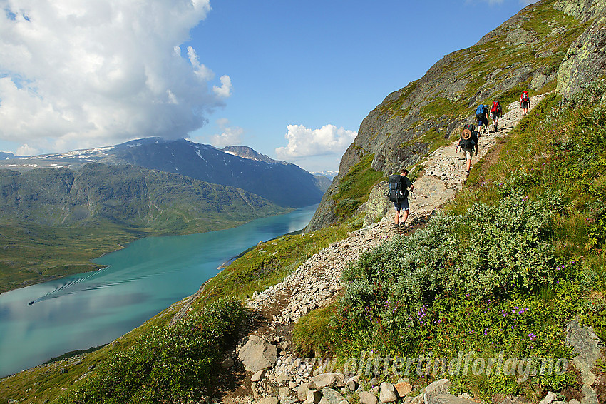 Fjellvandrer på tur over Besseggen. Her på vei opp fra Gjendesheim mot Veslfjellet. Nede til venstre ses Gjende med rutebåten Gjendinge på vei østover mot Gjendeosen. I bakgrunnen ses bl.a. Knutshøe, Bukkehåmåren, Høgdebrotet og Eggen.