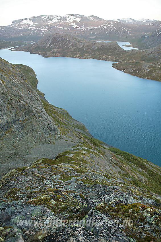 Utsikt fra Besseggen hvor man ser deler av Jo stigen ovenfra. Ruta følger ryggen midt på bildet et godt stykke nedover. Deretter trekker den til venstre i bunn av grusrenna til venstre, før den svinger til høyre nedenfor en skrent og kommer ut på en bratt, rødfarget klipperygg litt lenger nede til høyre. Deler av ruta er potensielt utsatt for steinsprang fra fjellveggen til venstre utenfor bildekanten.