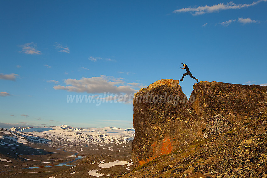 Sprang over luftig, kløftet, steinblokk på dalbrekket over Veodalen med bl.a. Surtningssue i bakgrunnen.