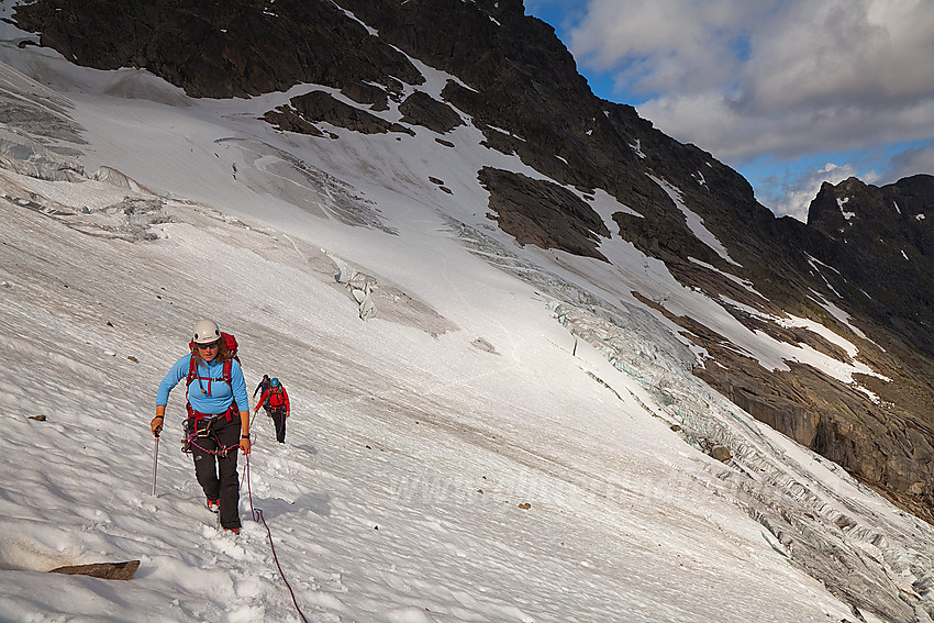 På vei over Slingsbybreen. Bak til høyre, omtrent på samme høyde som personene ses svaene man normalt krysser på vei til Maradalsryggen fra denne siden. Bakenfor disse svaene igjen ses et lengre snøfelt som skrår oppover. Ruta følger kanten av dette oppover fjellsiden.