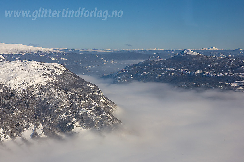 Vi har akkurat flydd over Heling og passerer over Vangsmjøse med utsikt nedover Valdres. Tåkeskyene ligger fortsatt tett over dalbunnen. Til høyre ses lett gjenkjennelige Hugakøllen med Rundemellen og Skarvemellen i det fjerne.