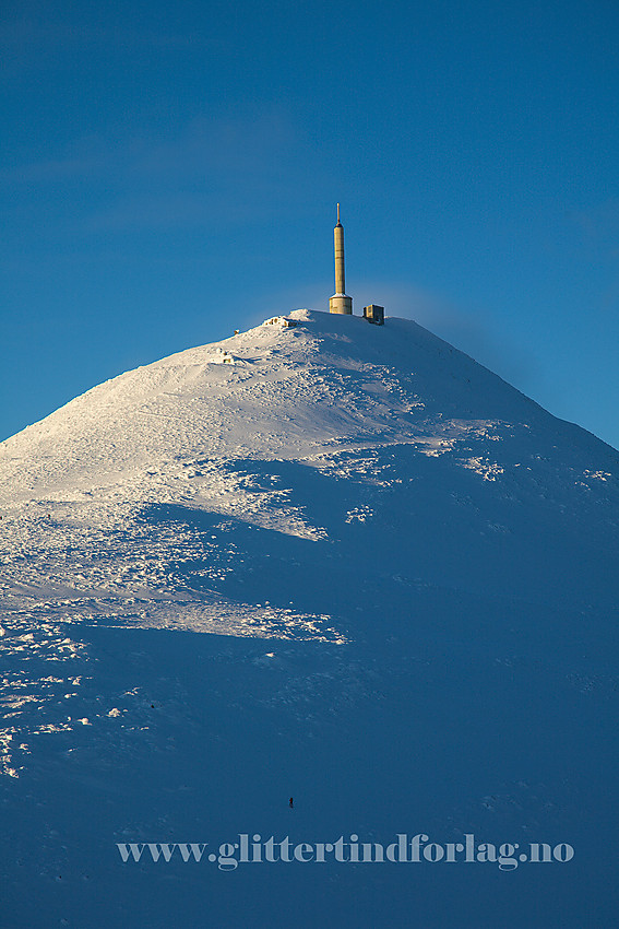 Gaustatoppen sett fra Gaustaråen i sør til sørøst. Selve toppen ligger litt bakenfor hytta og masta, utenfor synsfeltet. Nede i skyggen ser man en skiløper på vei opp mot toppen.