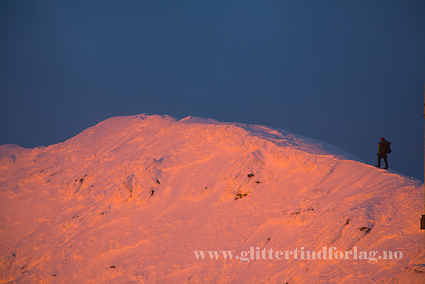 På vei bortetter toppryggen på Gaustatoppen en romjulskveld mens solnedgangen fyrer opp snøflanken. En vegg av skyer i bakgrunnen lager et mørkt bakteppe.