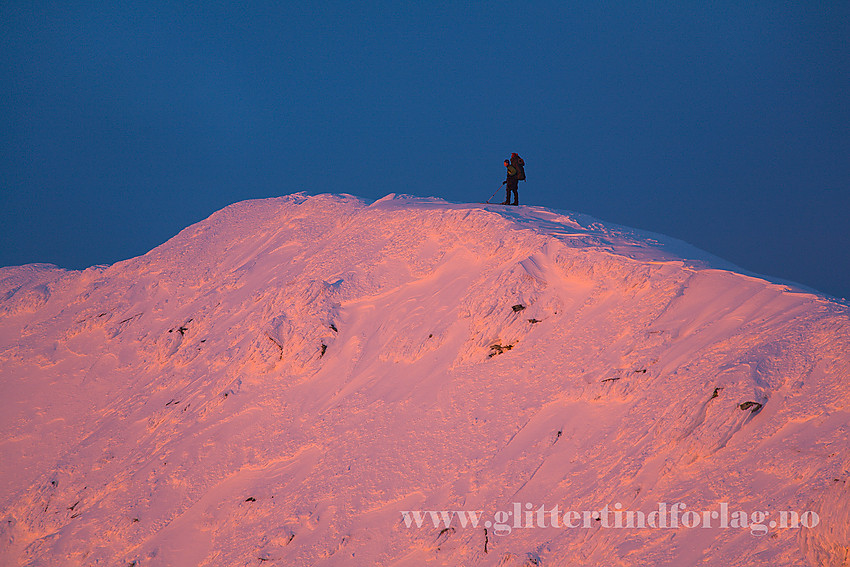 På vei bortetter toppryggen på Gaustatoppen en romjulskveld mens solnedgangen fyrer opp snøflanken. En vegg av skyer i bakgrunnen lager et mørkt bakteppe.