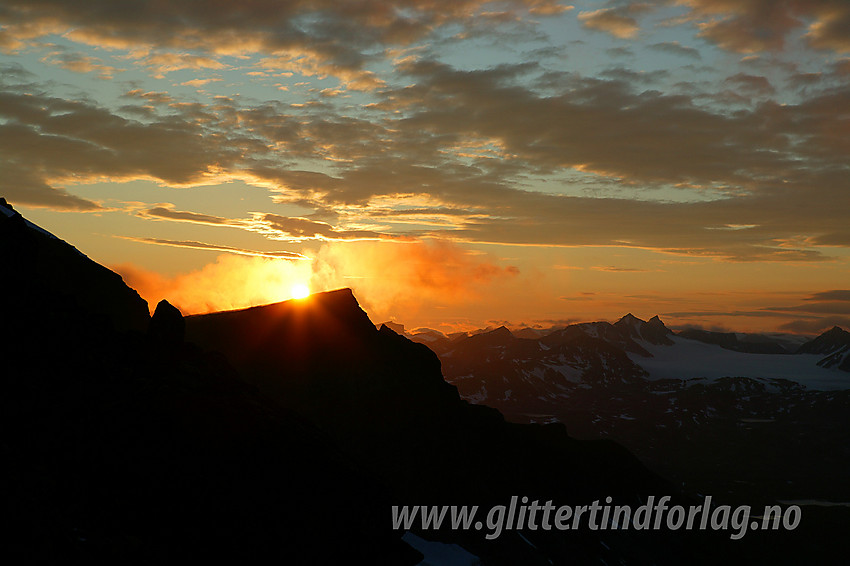 Fra Midtre Leirungstinden ved solnedgang med utsikt nordover. Sola går ned bak Nørdre Knutsholstinden (2185 moh) og gløder opp ei sky i oppløsning som siger over topplatået. Bak til høyre ses bl.a. Hellstugutindane og Vestre Memurubrean.