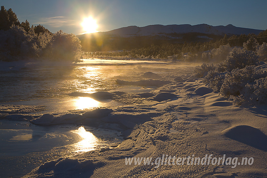 Ved Sjoa en bitende kald desembermorgen. Frostrøyken ligger over vannflata og fjellbjørkene er tunge av rimkrystaller. I bakgrunnen ses silhuetten av bl.a. Besstrondfjellet.