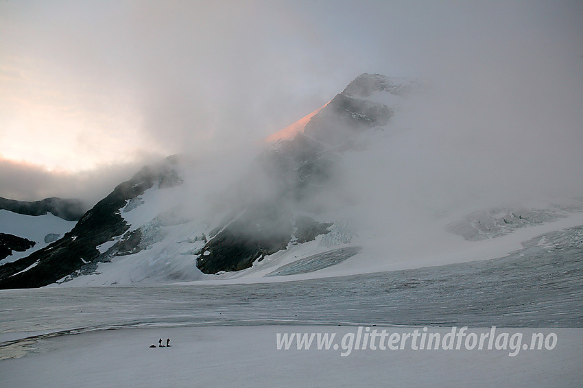 To små personer ute på breflata på Gjertvassbreen mens morgensola gløder på Gjertvasstinden (2351 moh). Rundt fjellryggene danser en rekke tåkeskyer.