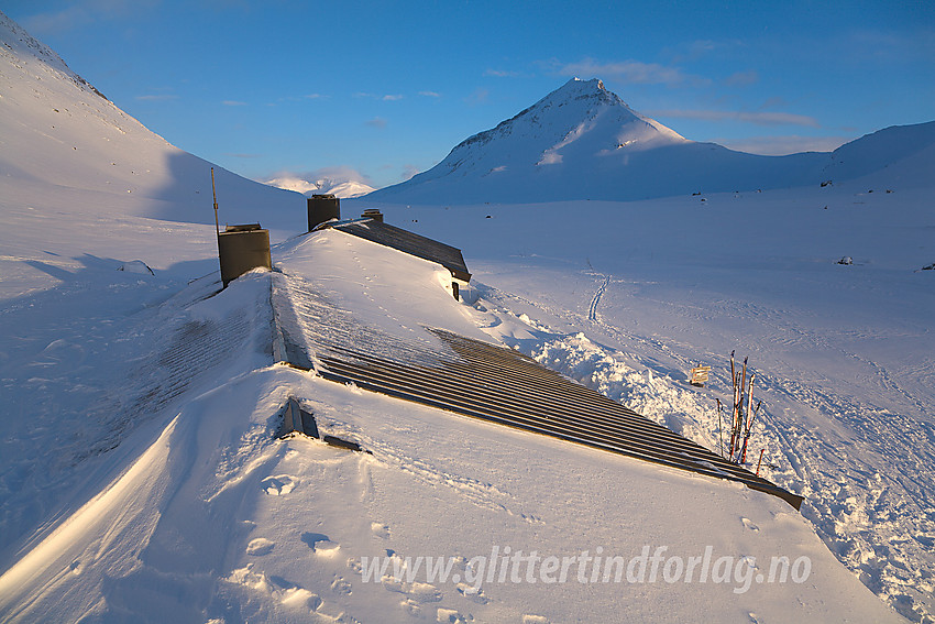 Taket på den nesten nedsnødde hovedhytte på Olavsbu med Snøholstinden (2141 moh) i bakgrunnen.